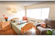 A bright and cozy bedroom in a senior living facility featuring a single bed with a green bedspread, a wooden nightstand with a lamp and flowers, a pink armchair near a large window with blinds, and a comfortable brown recliner chair with a colorful pillow.