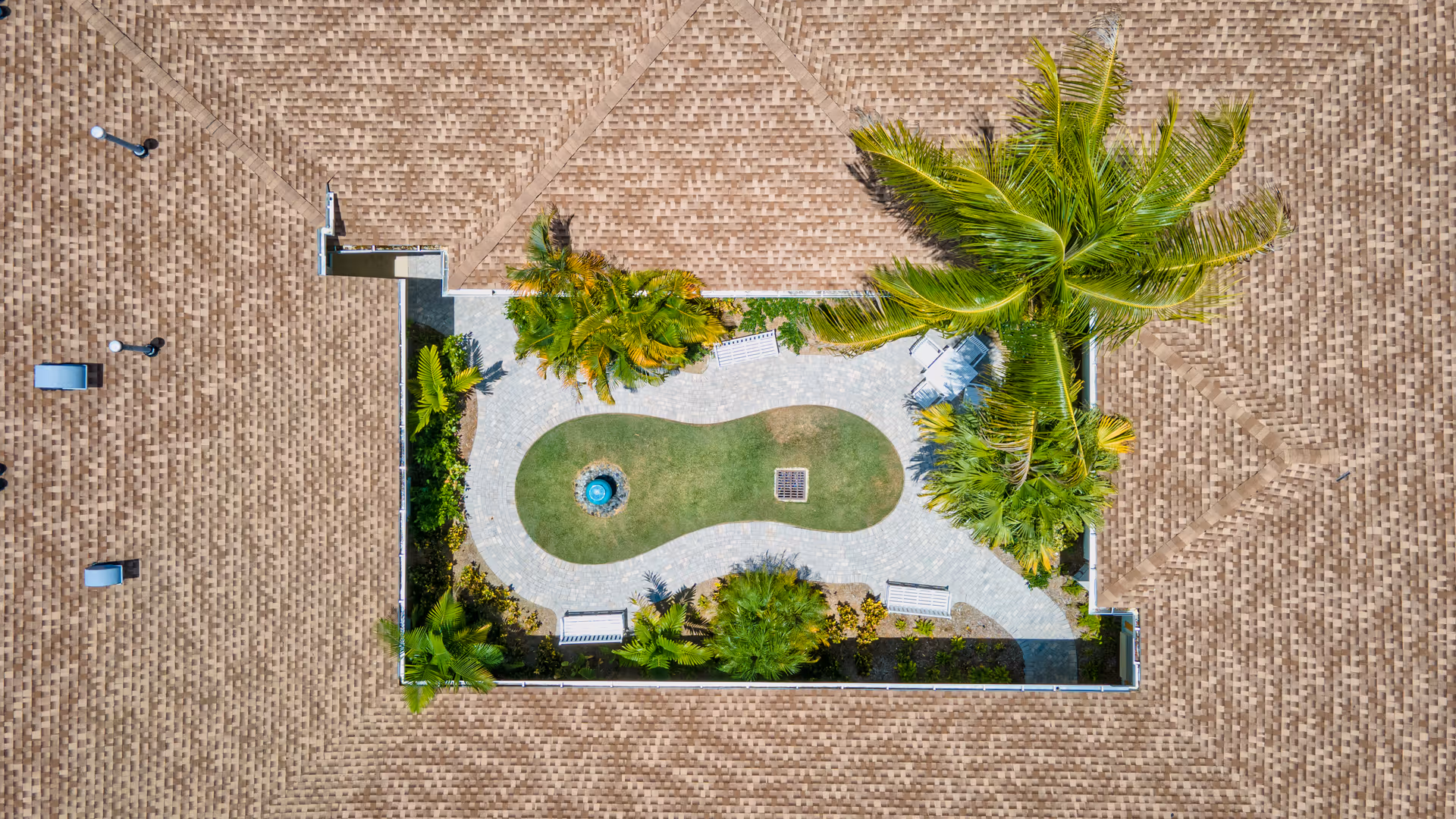 Aerial view of a rooftop garden area with a small green lawn, a circular water fountain, several palm trees, bushes, and white benches arranged along a curved paved pathway, surrounded by a brown shingled roof.