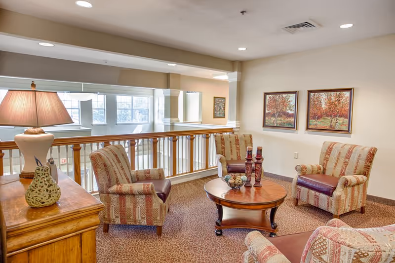 A cozy senior living common area with four upholstered armchairs arranged around a round wooden coffee table. The table holds decorative items including candles and a bowl with ornamental balls. The room has beige walls adorned with two framed paintings of autumn trees. A wooden side table with a lamp and a decorative pear sculpture is visible on the left. The space overlooks a lower level through a wooden railing, with large windows letting in natural light.