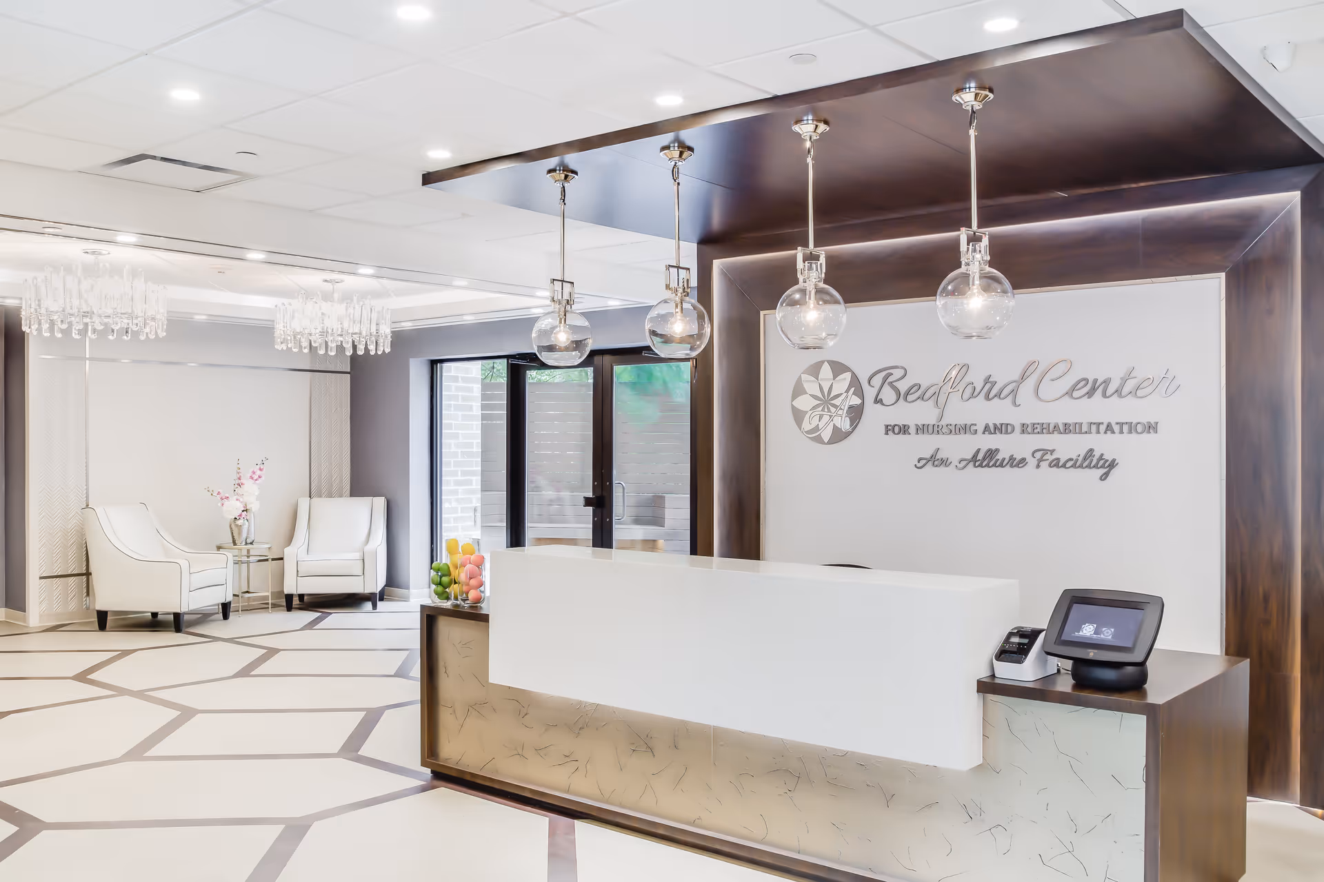 Reception area of Bedford Center for Nursing and Rehabilitation featuring a modern white reception desk with a dark wood backdrop, four hanging globe pendant lights, and a sign displaying the facility's name. The area includes two white armchairs with a small table holding a vase of flowers, geometric patterned flooring, and large glass doors leading outside.