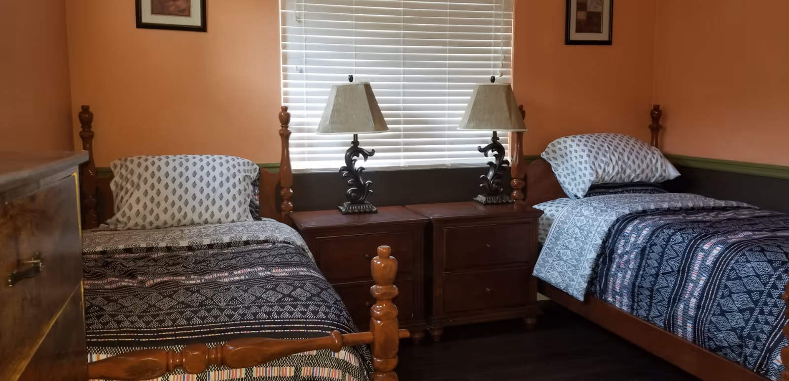 A bedroom with two single wooden beds, each with patterned bedding and pillows. Between the beds are two wooden nightstands with matching decorative lamps. A window with white blinds is centered behind the nightstands, and two framed pictures hang on the peach-colored walls above each bed.