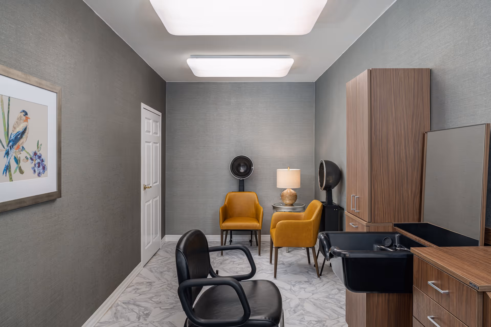 Interior view of a senior living facility hair salon with two mustard yellow chairs and a small round table with a lamp between them, a black salon chair in front of a wooden cabinet with a mirror and sink, gray textured walls, a framed bird painting on the left wall, and marble-patterned floor tiles.