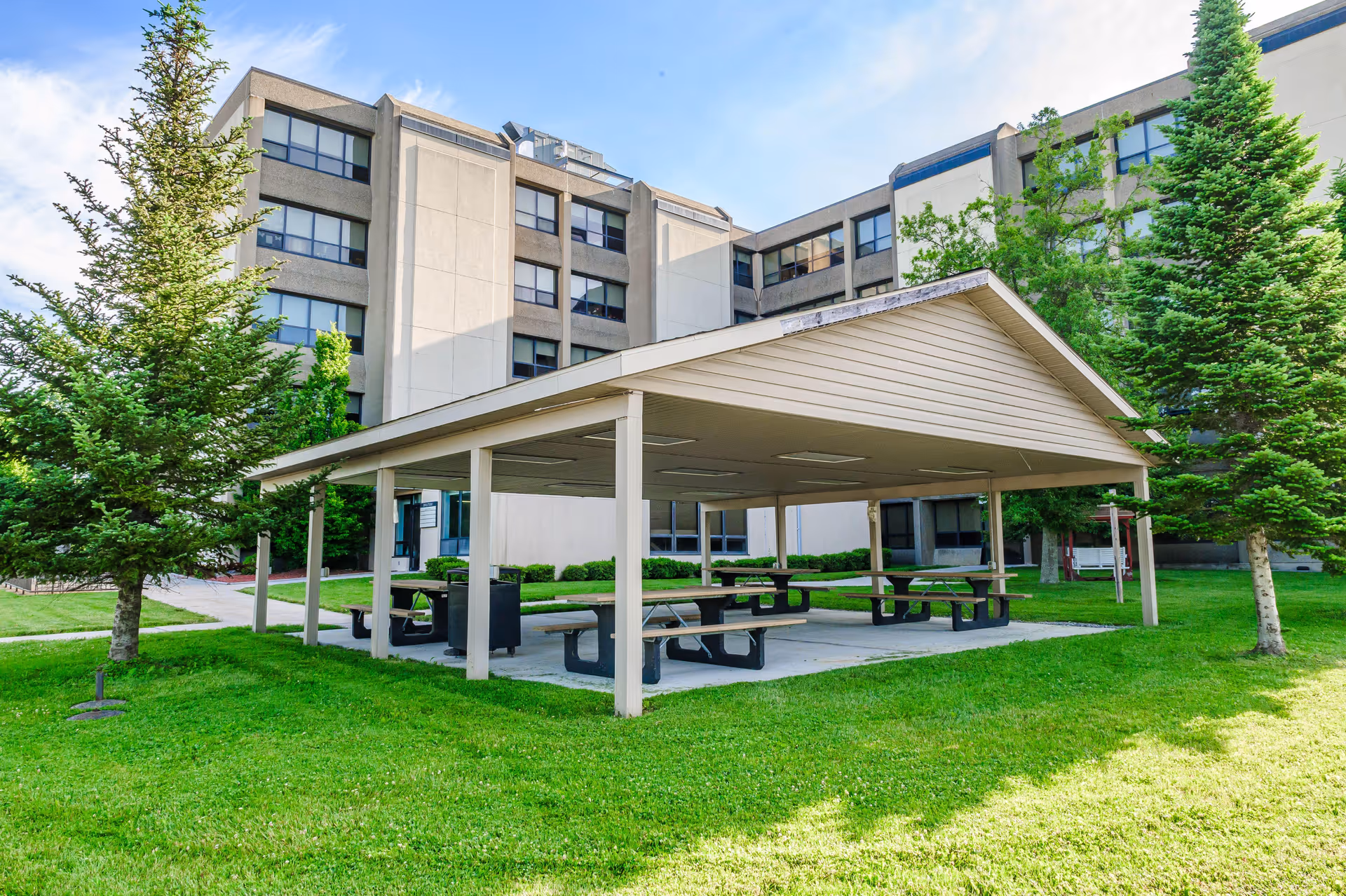 Covered outdoor picnic pavilion with several picnic tables on a grassy lawn in front of a multi-story health and rehab building.