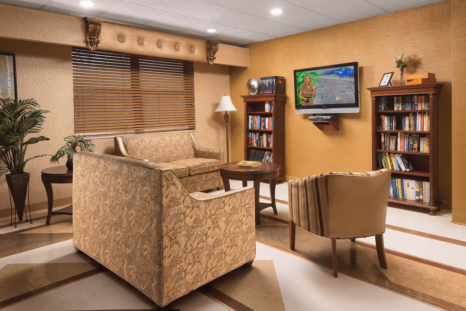 A cozy living room area with a floral-patterned sofa, a striped armchair, a round wooden coffee table, two wooden bookshelves filled with books, a floor lamp, and a wall-mounted TV displaying a weather forecast. The room has beige walls, a window with wooden blinds, and decorative plants.