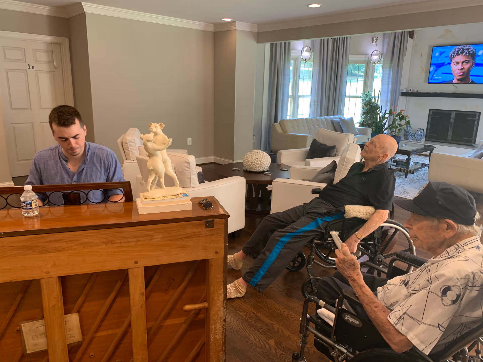 A young man playing a wooden piano in a living room area of a senior living facility. Two elderly men in wheelchairs are seated nearby, one looking towards the piano and the other holding a remote control. The room has white armchairs, a coffee table, a fireplace, and a TV mounted on the wall showing a person's face.