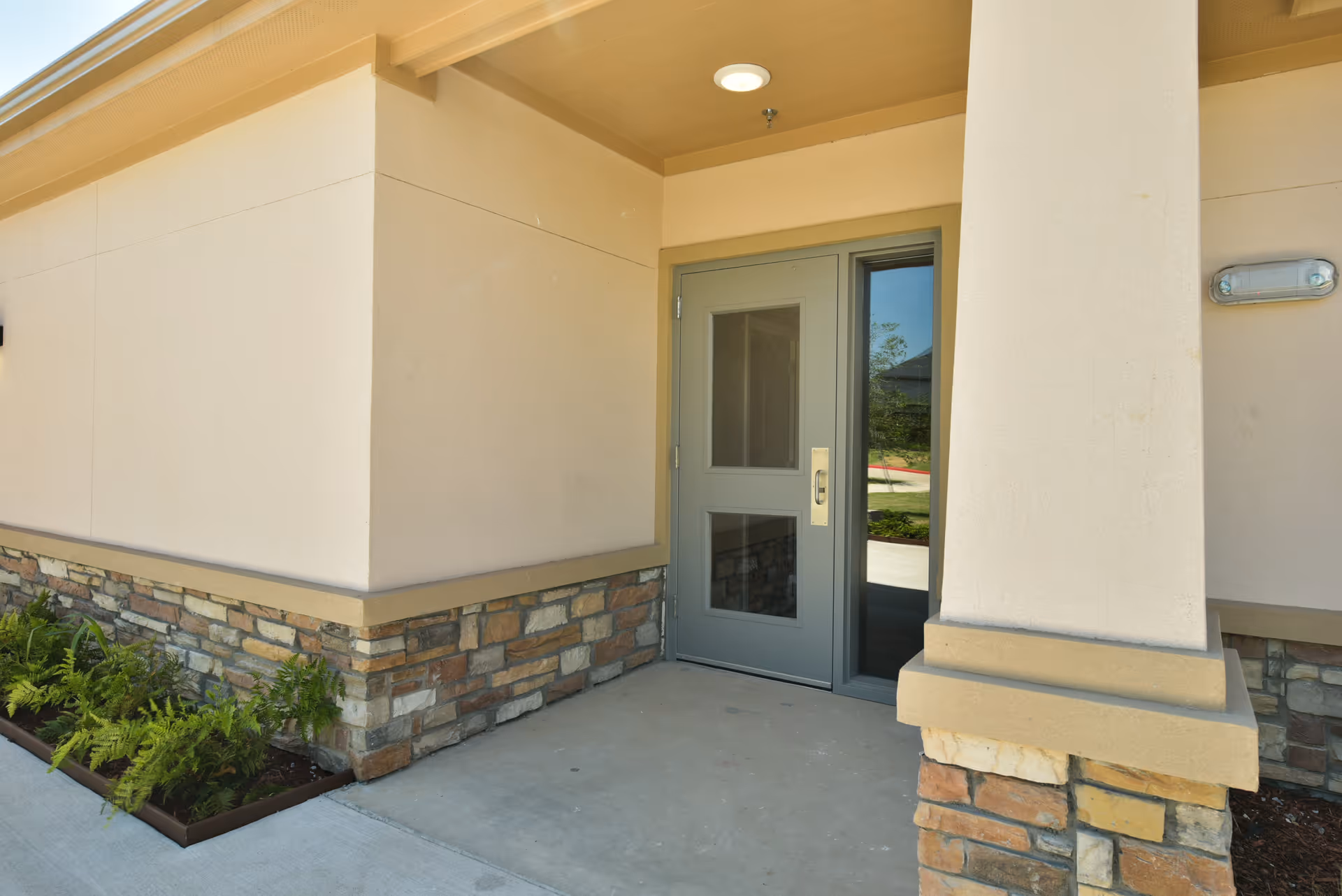 Exterior entrance of a building with a beige wall, stone accents at the base, a gray door with glass panels, and a small garden bed with green plants along the side.