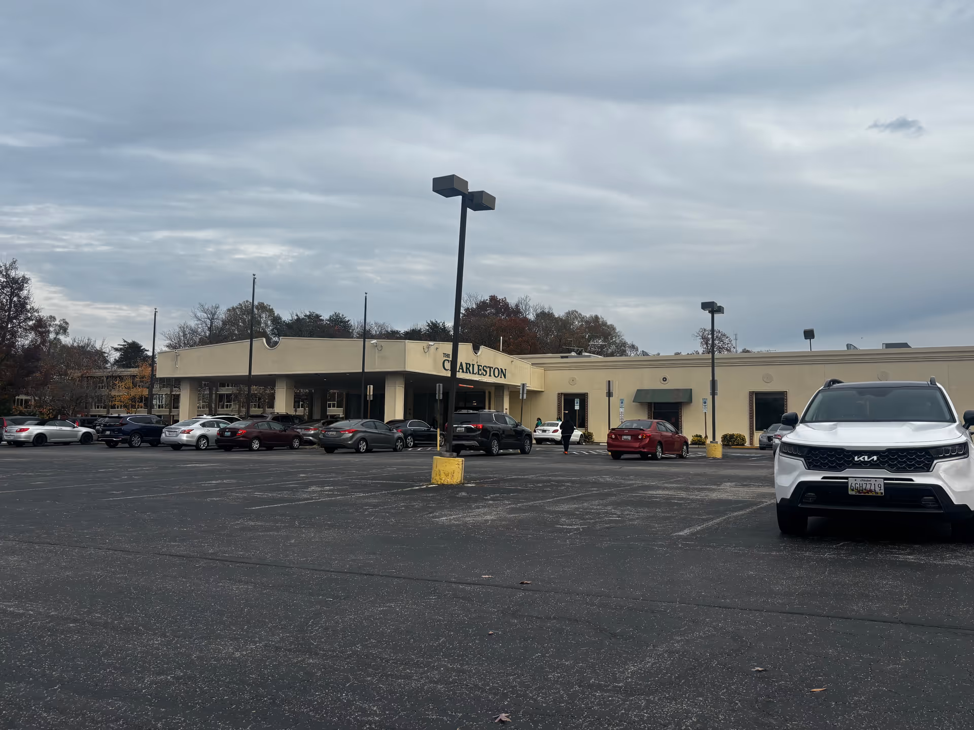 Exterior view of The Charleston Senior Community building with a large parking lot in front. Several cars are parked, and a person is walking towards the entrance. The sky is cloudy.