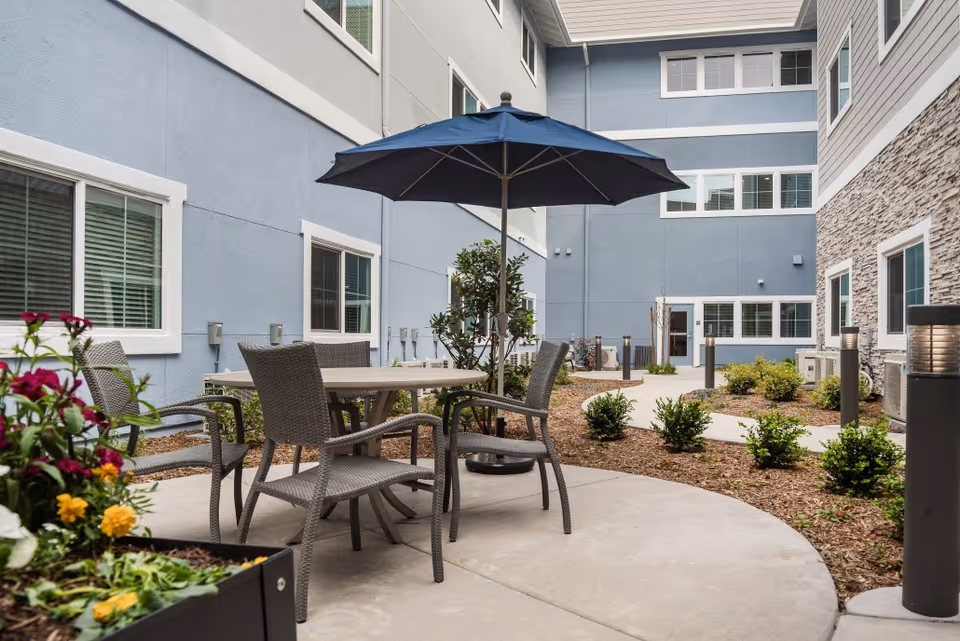 Courtyard patio with a round table, wicker chairs and a blue umbrella surrounded by landscaping and multi-story building façades.