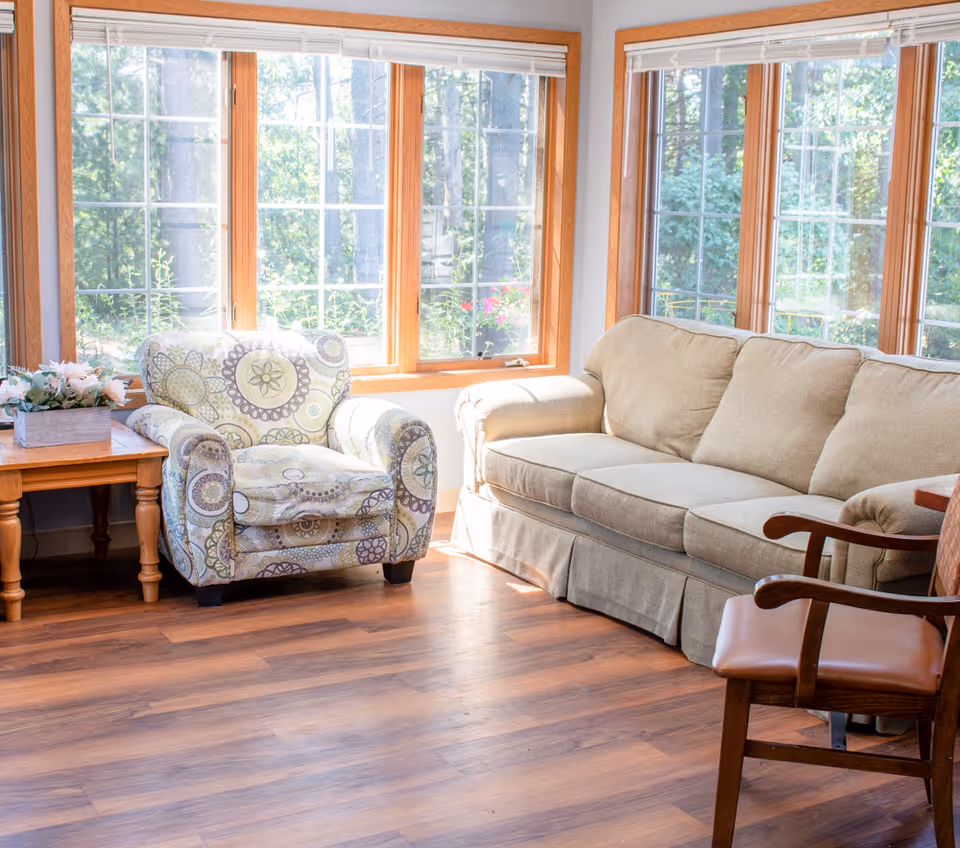 Sunlit living room with a patterned armchair, beige sofa, wooden side table with flowers, and large mullioned windows showing greenery.