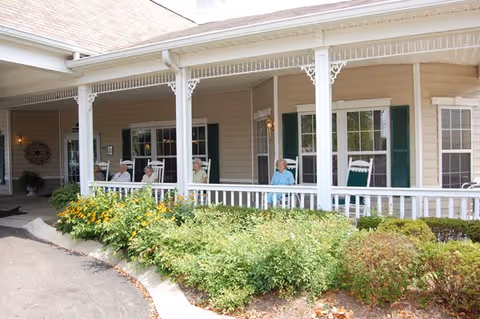 A covered porch area of a senior living facility with several elderly people sitting on white rocking chairs. The porch has white railings and decorative trim, with green shrubs and yellow flowers in front. The building exterior is beige with green shutters on the windows.