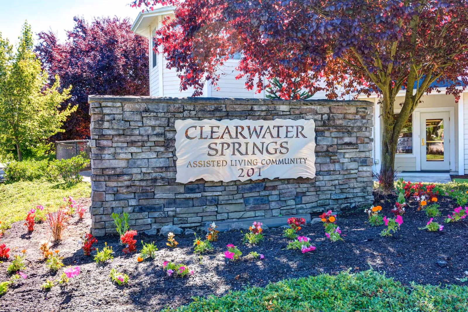 Stone sign for Clearwater Springs Assisted Living Community with the number 201, surrounded by colorful flowers and trees, with part of a white building visible in the background.