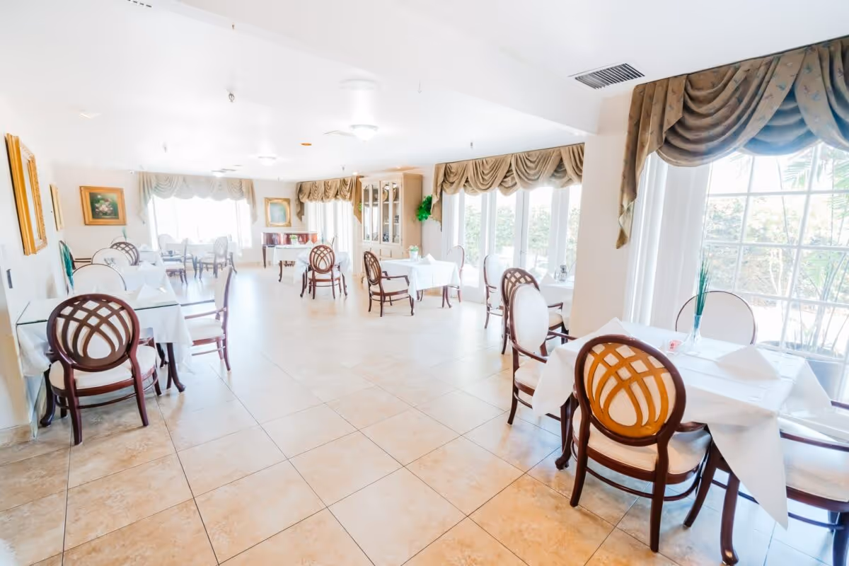 Bright dining room with multiple tables covered in white tablecloths, each surrounded by elegant wooden chairs with cushioned seats. Large windows with draped curtains allow natural light to fill the room. The floor is tiled, and paintings hang on the walls.