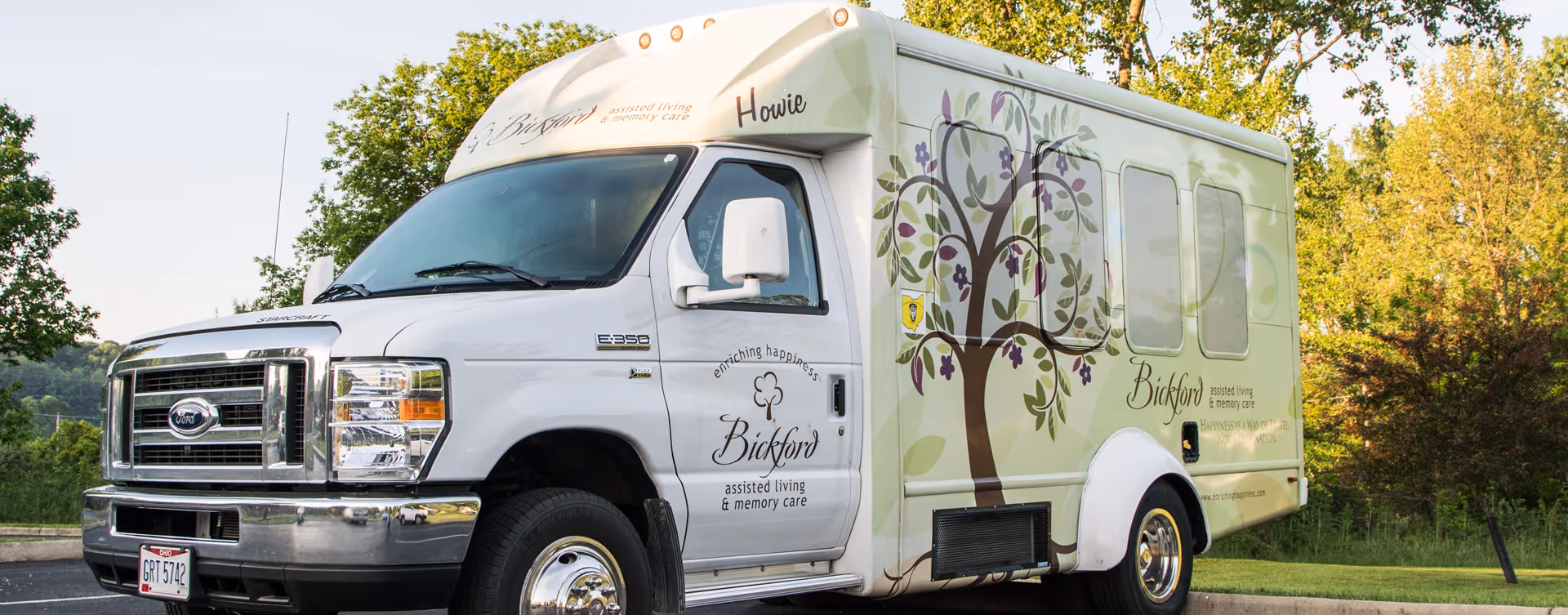 A white Ford shuttle bus with floral and tree graphics parked outdoors near greenery. The bus has branding for Bickford assisted living & memory care, including the phrases 'enriching happiness' and 'Happiness is a way of travel, not a destination.'