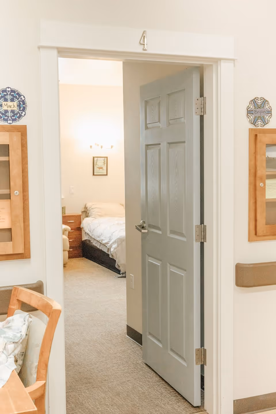 View through an open door labeled number 4 into a bedroom with a bed, nightstand, and wall light fixture. The hallway outside the door has a chair and bulletin boards on the walls with name tags 'Mack' and 'Bryan'.