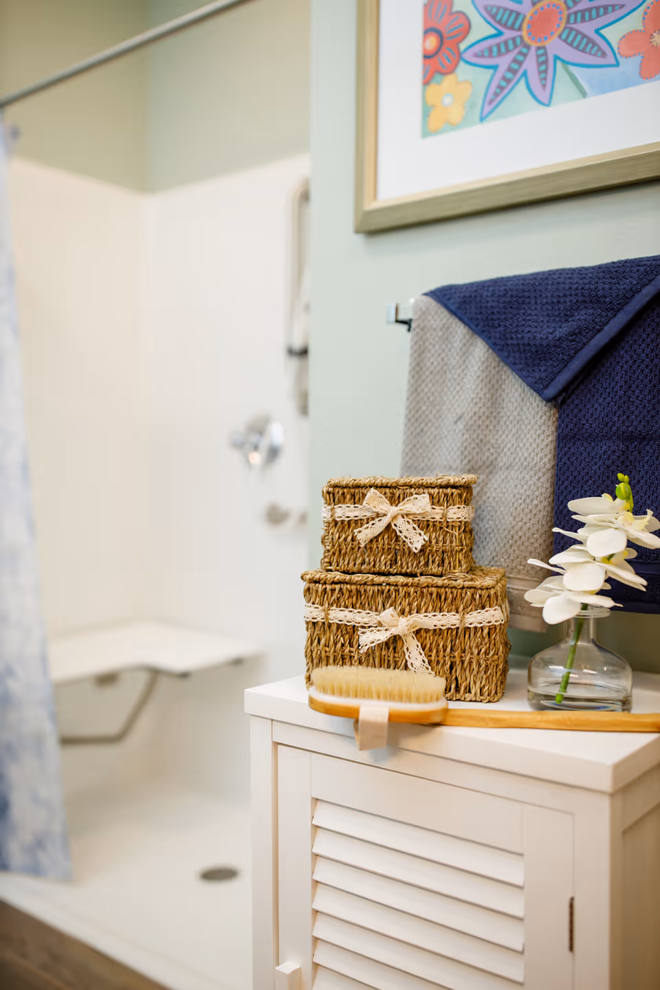 A bathroom corner featuring a white cabinet with two woven baskets tied with lace ribbons, a wooden brush, a glass vase with white flowers, and folded towels hanging on a rack. In the background, there is a shower area with a built-in seat and a floral framed artwork on the wall.