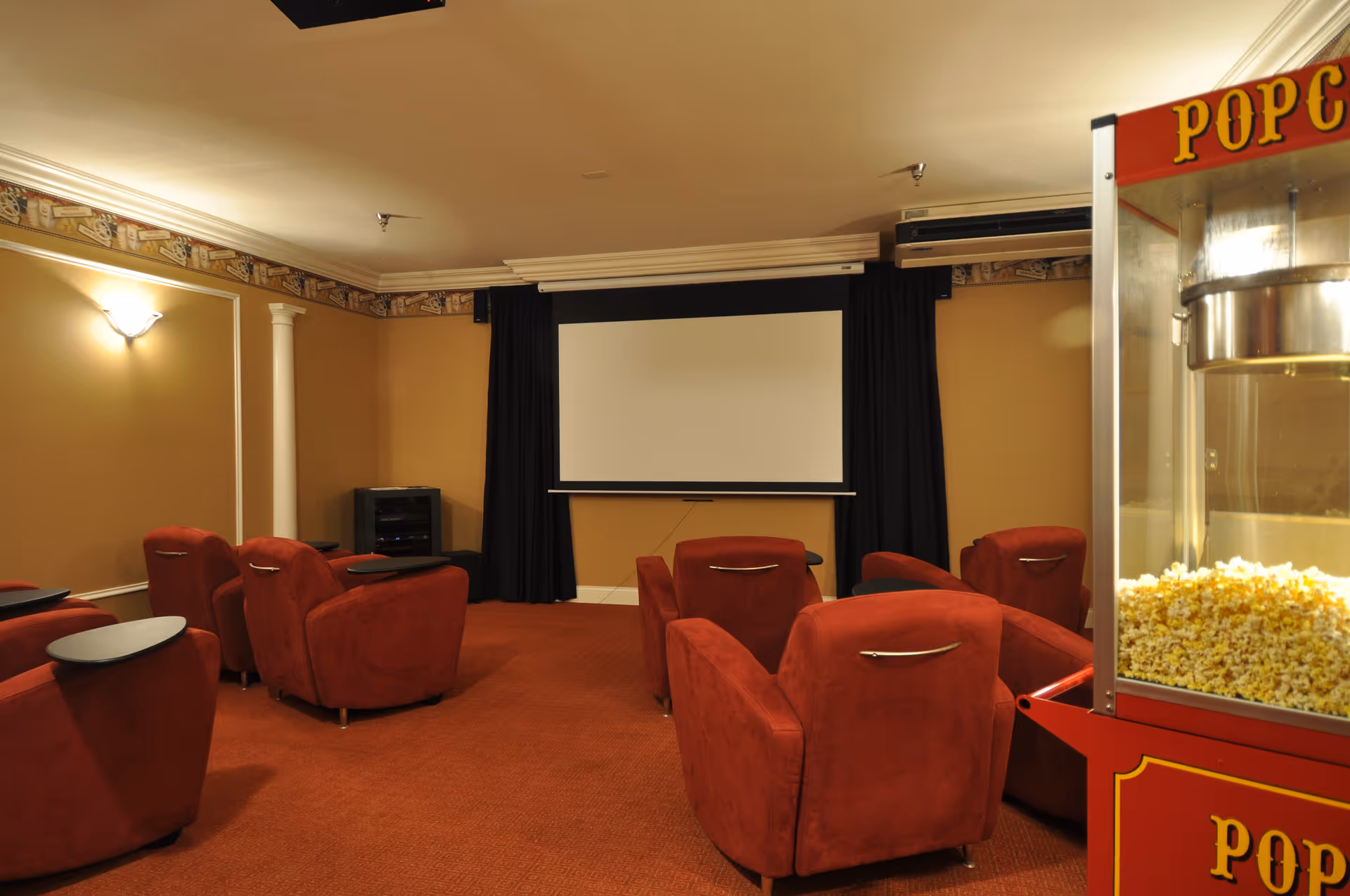 Small screening room with red upholstered recliners facing a projection screen and a popcorn machine at right.