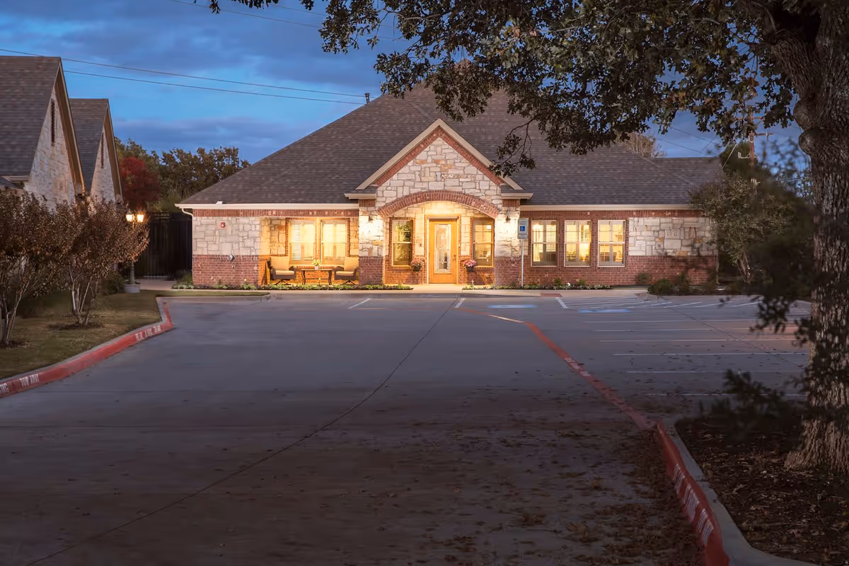Exterior view of a single-story brick and stone building with a peaked roof, warmly lit from inside during dusk. The building is surrounded by a parking lot with marked spaces and bordered by trees and shrubs.