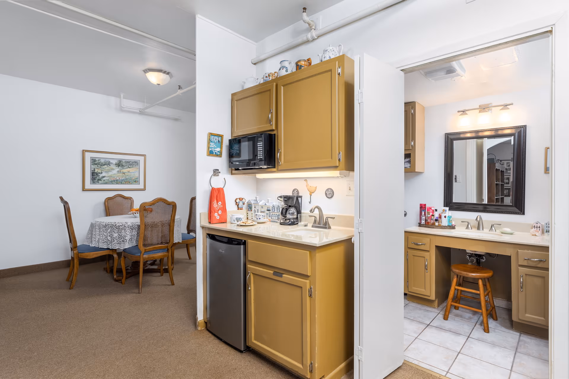 Interior view of a senior living facility showing a small kitchenette with mustard-colored cabinets, a microwave, coffee maker, and mini fridge. To the right, there is a bathroom vanity area with a large mirror, sink, and wooden stool. In the background, a dining area with a round table covered with a lace tablecloth and four wooden chairs is visible. The walls are white, and the floor is carpeted in the dining area and tiled in the bathroom.