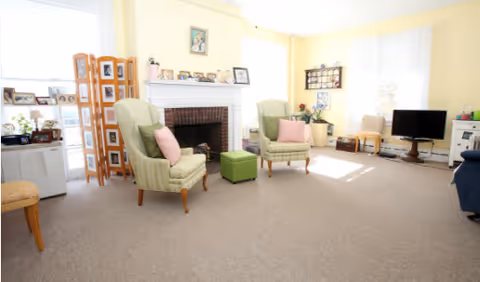 A bright living room with beige carpet, two green armchairs with pink pillows, a small green ottoman, a white fireplace with framed photos and decorations on the mantel, a TV on a stand, and large windows with white curtains letting in natural light.