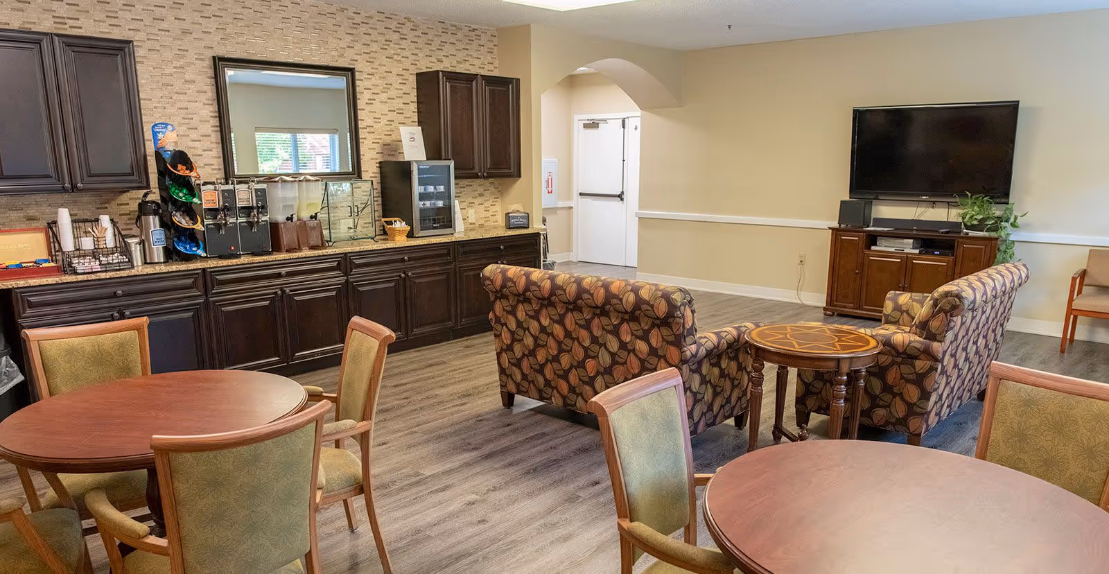 A common area in a senior living facility featuring round wooden tables with chairs, two patterned armchairs around a small round wooden table, a large flat-screen TV mounted on the wall above a wooden cabinet, and a counter with coffee and beverage dispensers along the back wall.