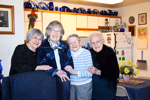 Four elderly women standing closely together in a kitchen area, smiling and posing for the photo. The kitchen has white cabinets with blue decorative items on top, a refrigerator with magnets, and a small dining table with a vase of yellow flowers.