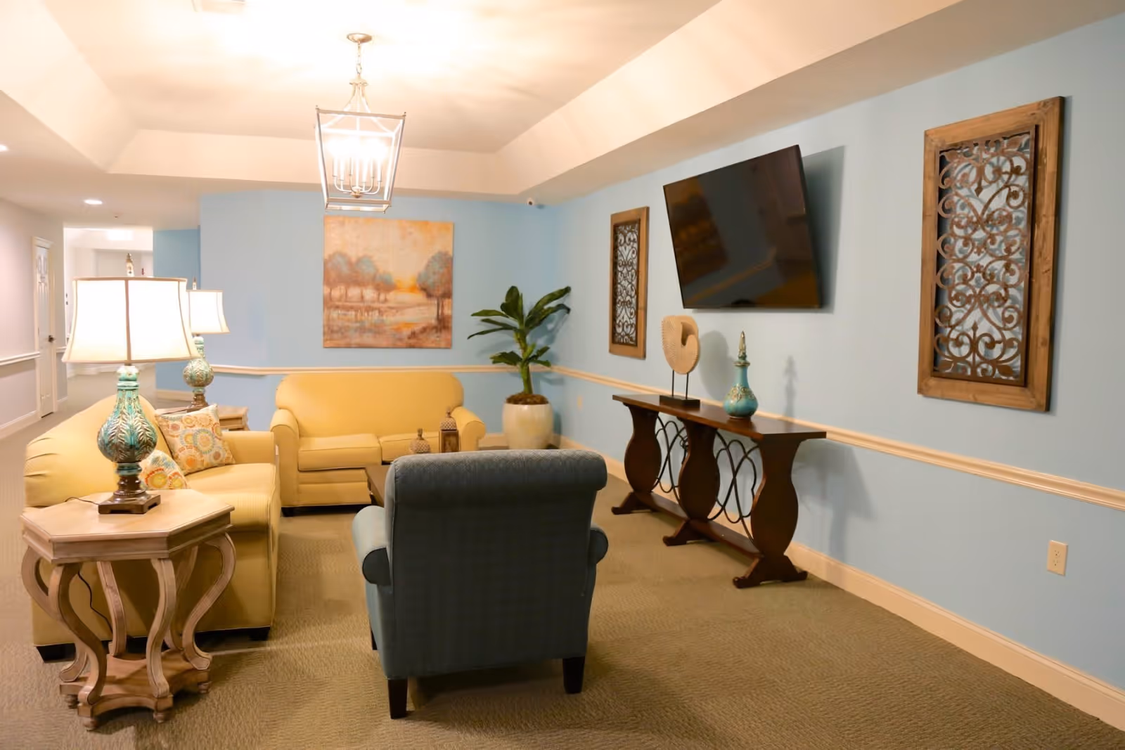 A cozy senior living room with light blue walls and beige carpet. The room features a yellow sofa, a matching loveseat, and a blue armchair arranged around a wooden side table with a decorative lamp. On the wall, there is a flat-screen TV mounted above a wooden console table with decorative items. Two framed ornamental wall hangings and a landscape painting are also visible. A potted plant is placed in the corner, and the room is softly lit by ceiling and table lamps.