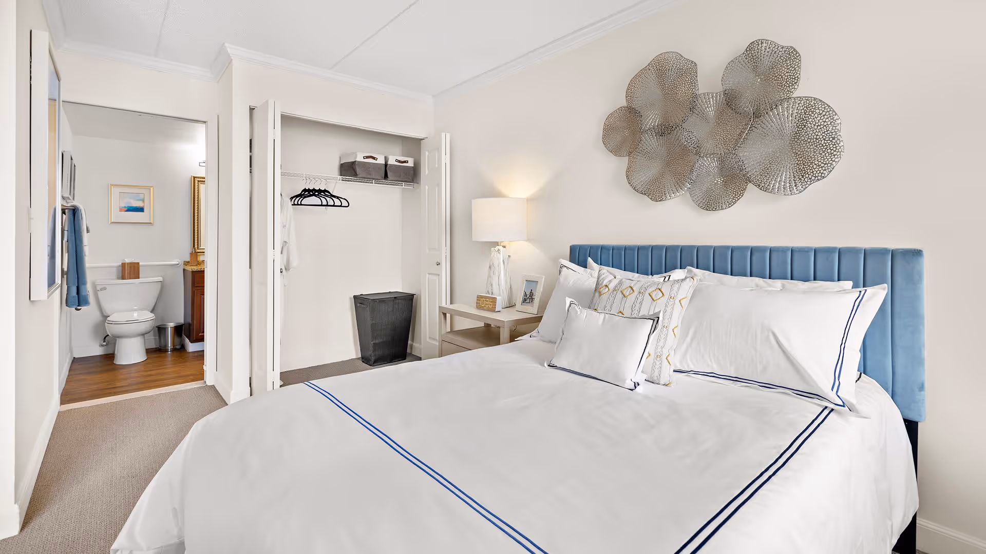 A neatly made bed with white linens and blue trim in a bedroom. The bed has a blue upholstered headboard and several pillows. To the right of the bed is a nightstand with a lamp and framed photo. On the wall above the bed is a decorative metal wall art piece. To the left, there is an open closet with hangers and storage bins, and an open door leading to a bathroom with a toilet and wooden floor.
