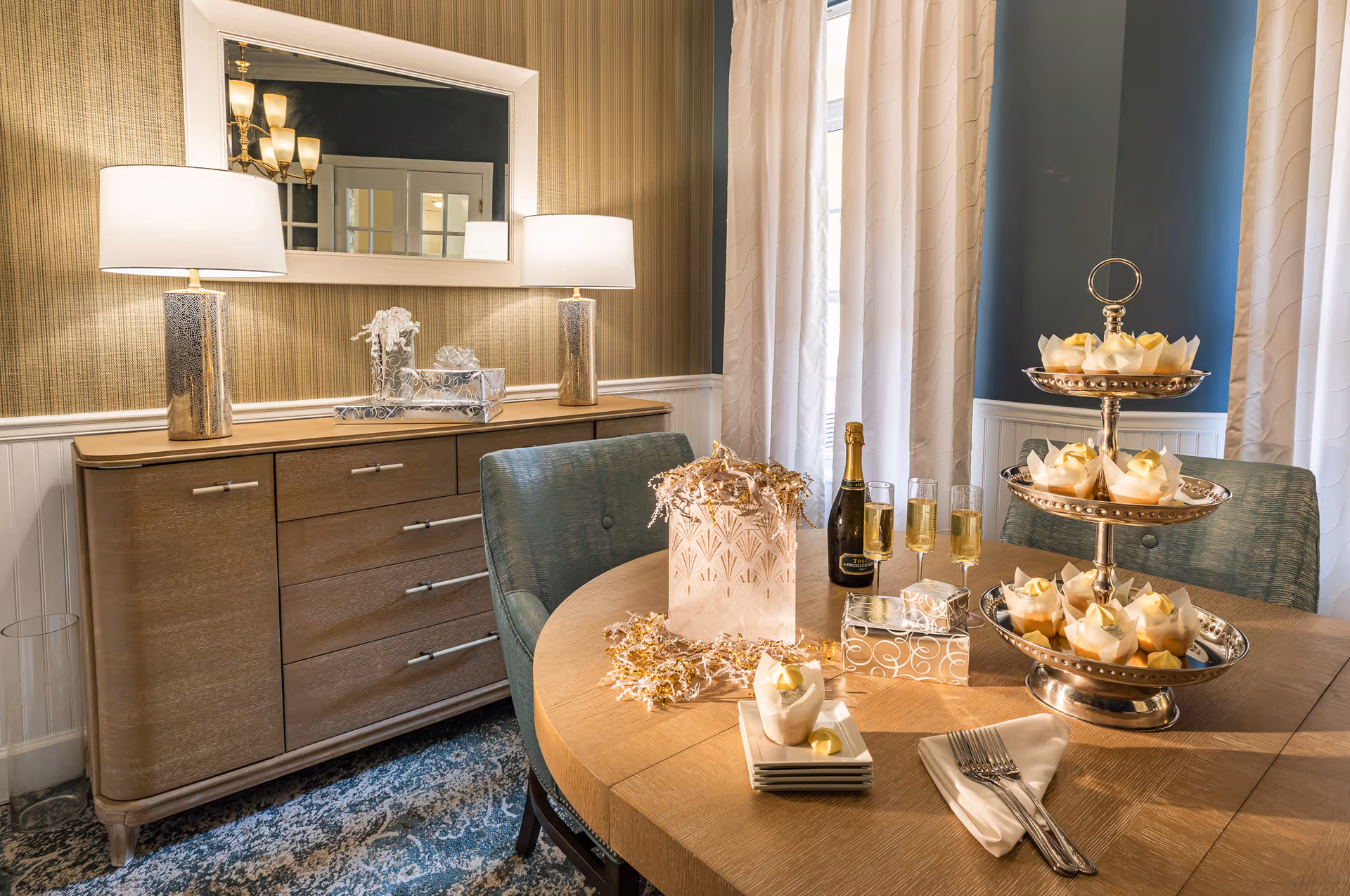 A warmly lit dining room with a wooden table set for a small gathering. On the table, there is a three-tiered silver tray holding cupcakes with white frosting, a bottle of champagne with four filled champagne flutes, a stack of small plates with a napkin and forks, and a decorative gift bag with gold and white shredded paper. Behind the table is a wooden sideboard with two lit lamps and a large mirror reflecting a chandelier and French doors. The room has beige striped wallpaper, white wainscoting, and light-colored curtains.