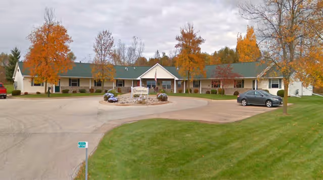 Single-story building with a green roof and beige exterior, surrounded by autumn-colored trees and a circular driveway with a sign in front. A car is parked on the right side and a red vehicle is partially visible on the left.