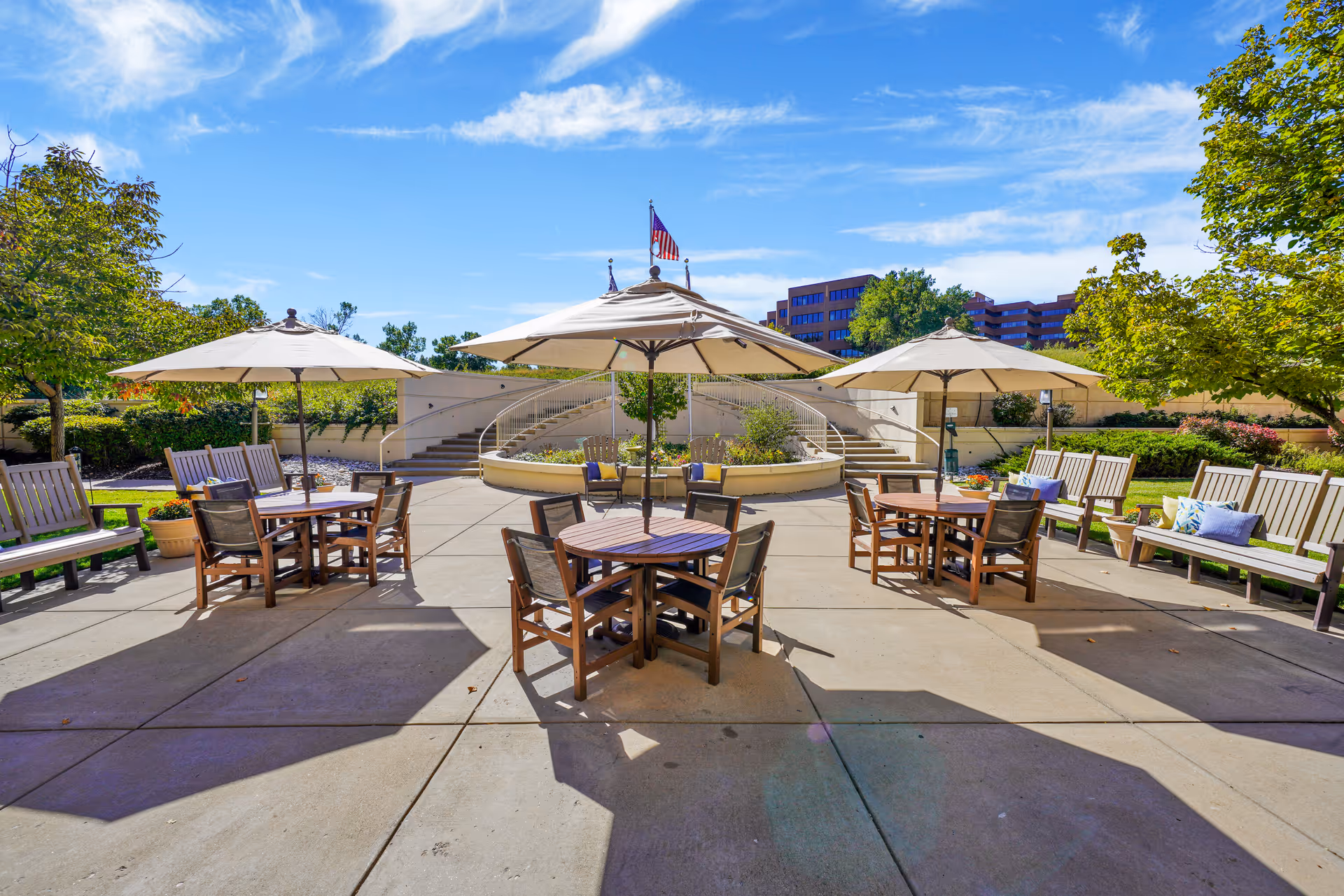 Outdoor patio area with round wooden tables and chairs under large beige umbrellas, surrounded by wooden benches with colorful cushions. There are trees and greenery around the patio, with a curved staircase and an American flag in the background under a blue sky with some clouds.
