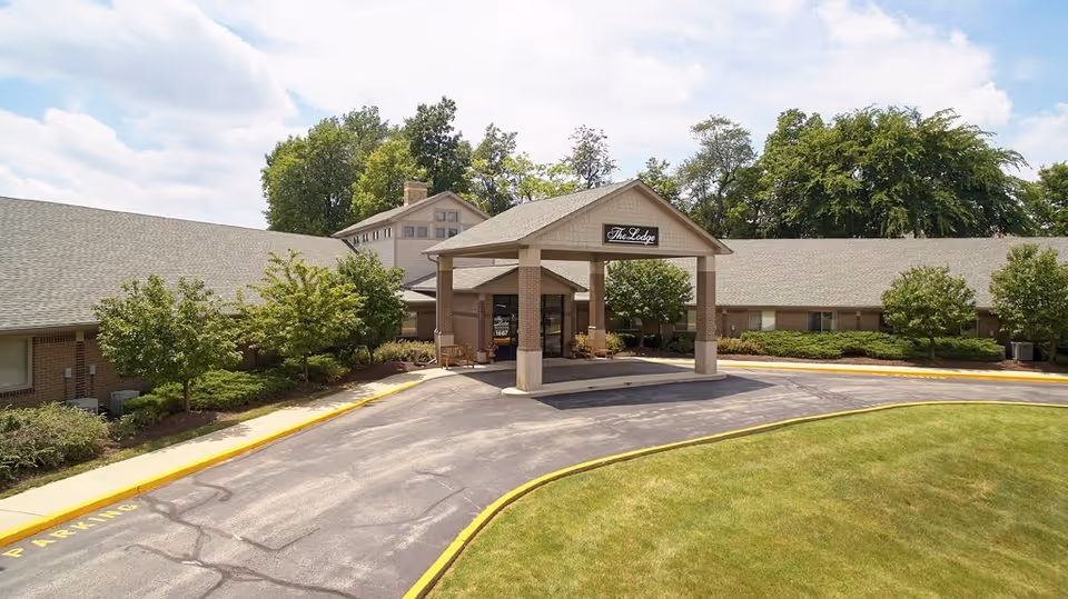 Exterior view of a single-story building with a covered entrance labeled 'The Lodge'. The building is surrounded by trees, shrubs, and a well-maintained lawn under a partly cloudy sky.