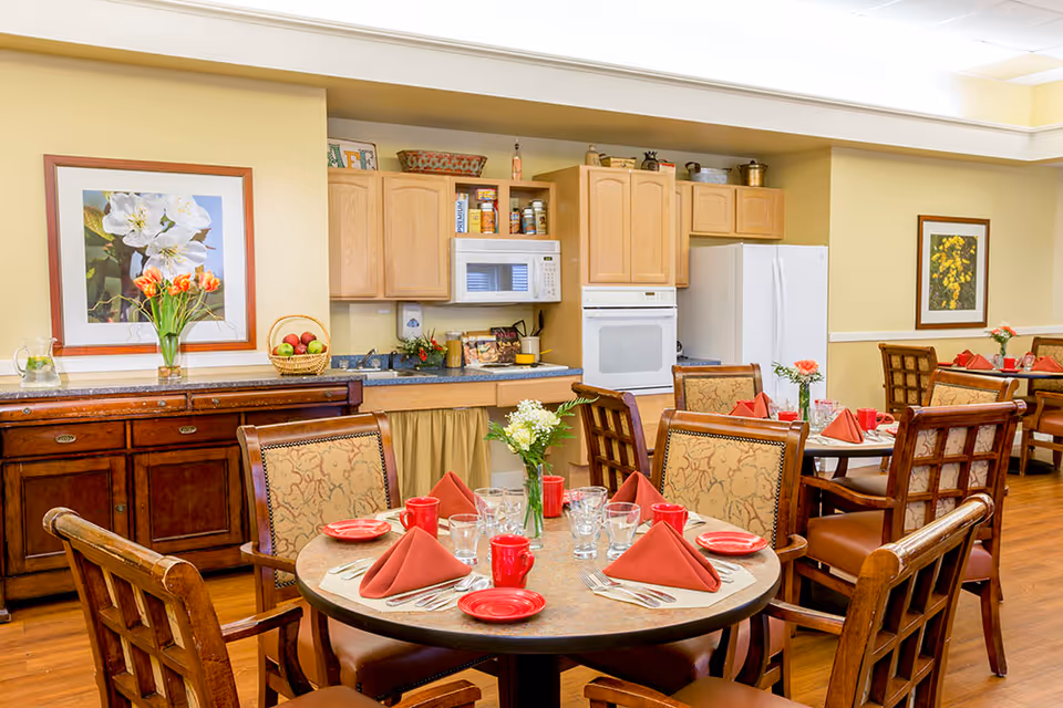 A dining area in a senior living facility with round tables set for a meal. Each table has red napkins, red plates, glasses, and silverware. The chairs have patterned upholstery. In the background, there is a kitchen area with wooden cabinets, a microwave, an oven, and a refrigerator. The walls are decorated with framed floral artwork and there are vases with flowers on the tables and counter.