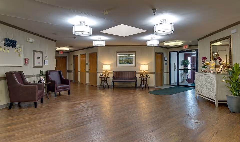 A spacious and well-lit nursing center lobby with wooden flooring, two brown leather armchairs on the left, a bench with two lamps on side tables against the back wall, and a white cabinet with decorative items and plants on the right. The entrance door is visible at the back right, and there are several wooden doors along the back wall. Ceiling lights and a skylight illuminate the area.