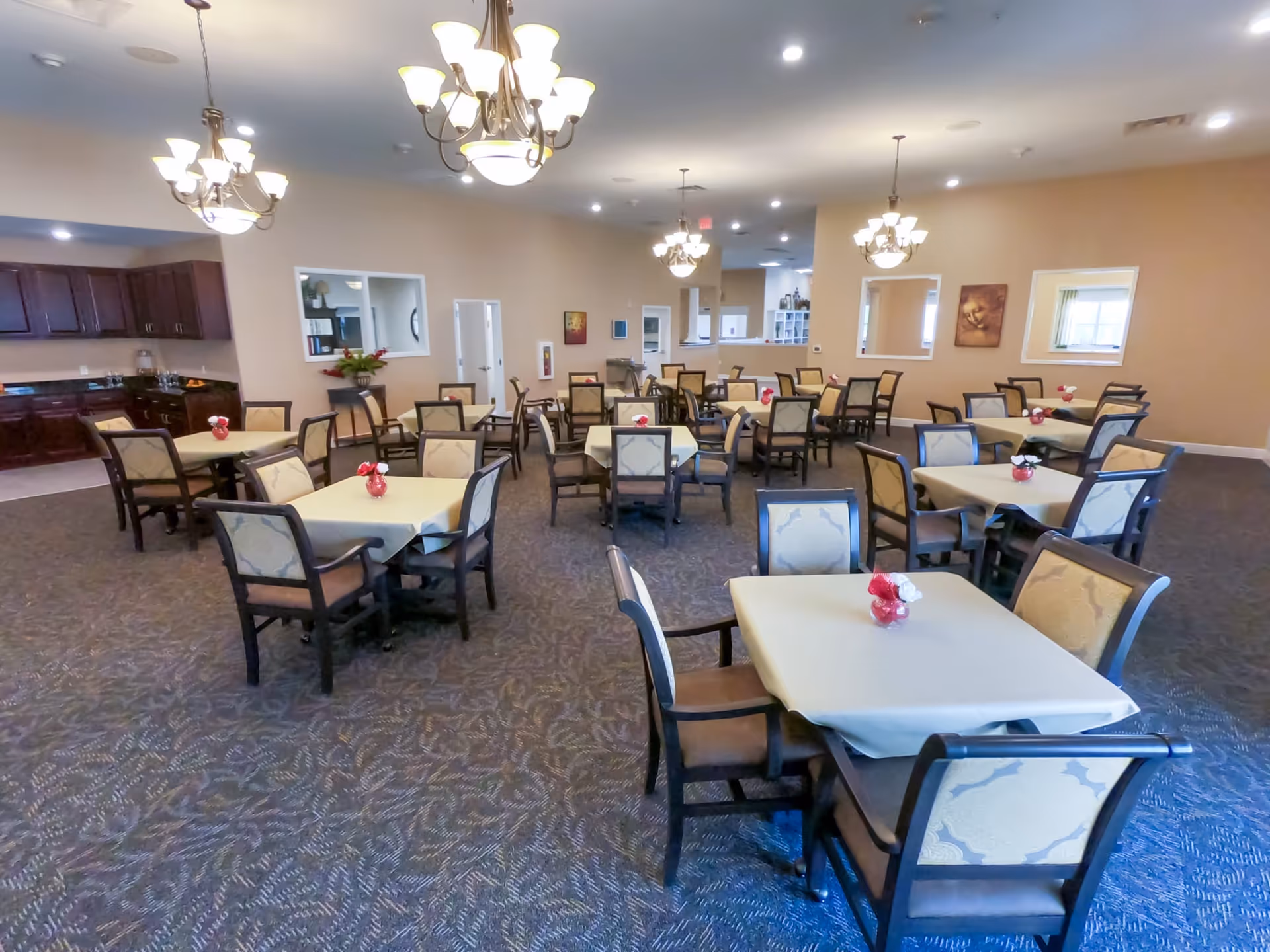 A spacious dining room with multiple square tables covered with beige tablecloths, each surrounded by four upholstered chairs. Small red vases with white and red flowers decorate each table. The room has beige walls, carpeted floors, and several chandeliers hanging from the ceiling. There is a kitchen area visible in the background with dark wood cabinets and a countertop.