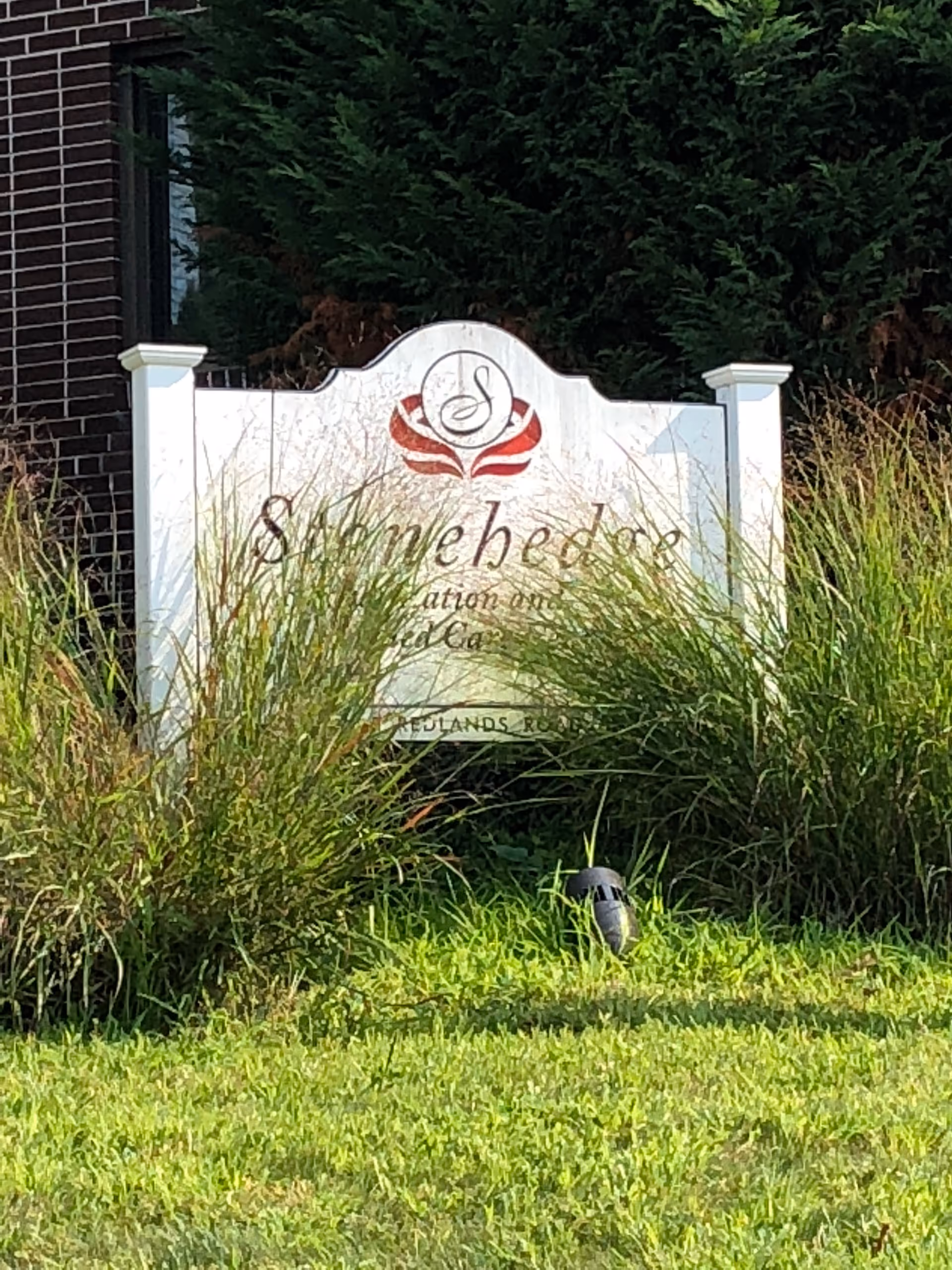 White entrance sign for Stonehedge Rehabilitation and Skilled Care partially obscured by tall grass in front of a brick building and greenery.