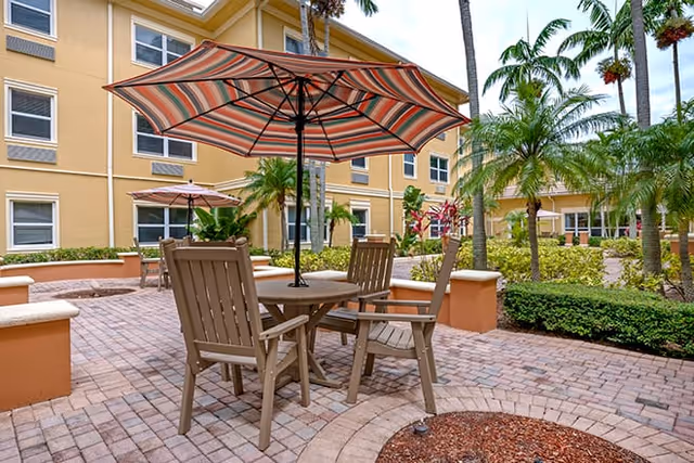 Outdoor courtyard with a round table, striped umbrella and wooden chairs on a brick patio surrounded by palm trees and a yellow apartment building.