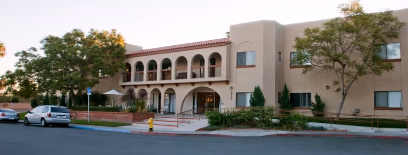 Exterior view of a two-story senior living facility building with beige stucco walls and a red tile roof. The building features arched balconies on the second floor and several windows. There are trees and shrubs around the entrance, a wheelchair ramp, a fire hydrant, and parked cars along the street in front.