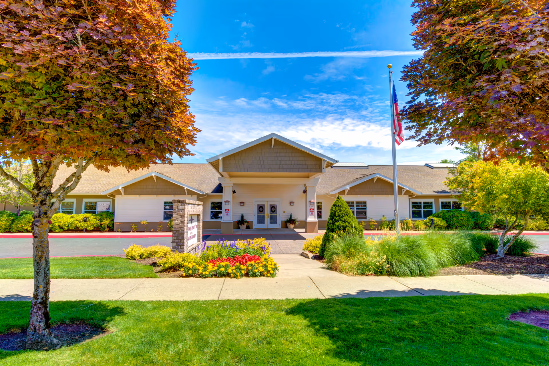 Front exterior view of Rosewood Memory Care facility with a well-maintained garden, colorful flowers, green shrubs, and two trees with reddish leaves framing the entrance. The building has a beige facade with a peaked roof and a flagpole with an American flag in front.