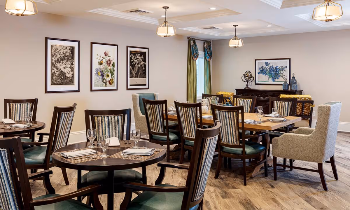 A well-lit dining room with multiple wooden tables and chairs arranged neatly. The chairs have striped upholstery and green cushions. The tables are set with placemats, napkins, and glassware. The walls are decorated with framed floral artwork, and there are pendant lights hanging from the ceiling. A window with green curtains and a sideboard with decorative items are visible in the background.