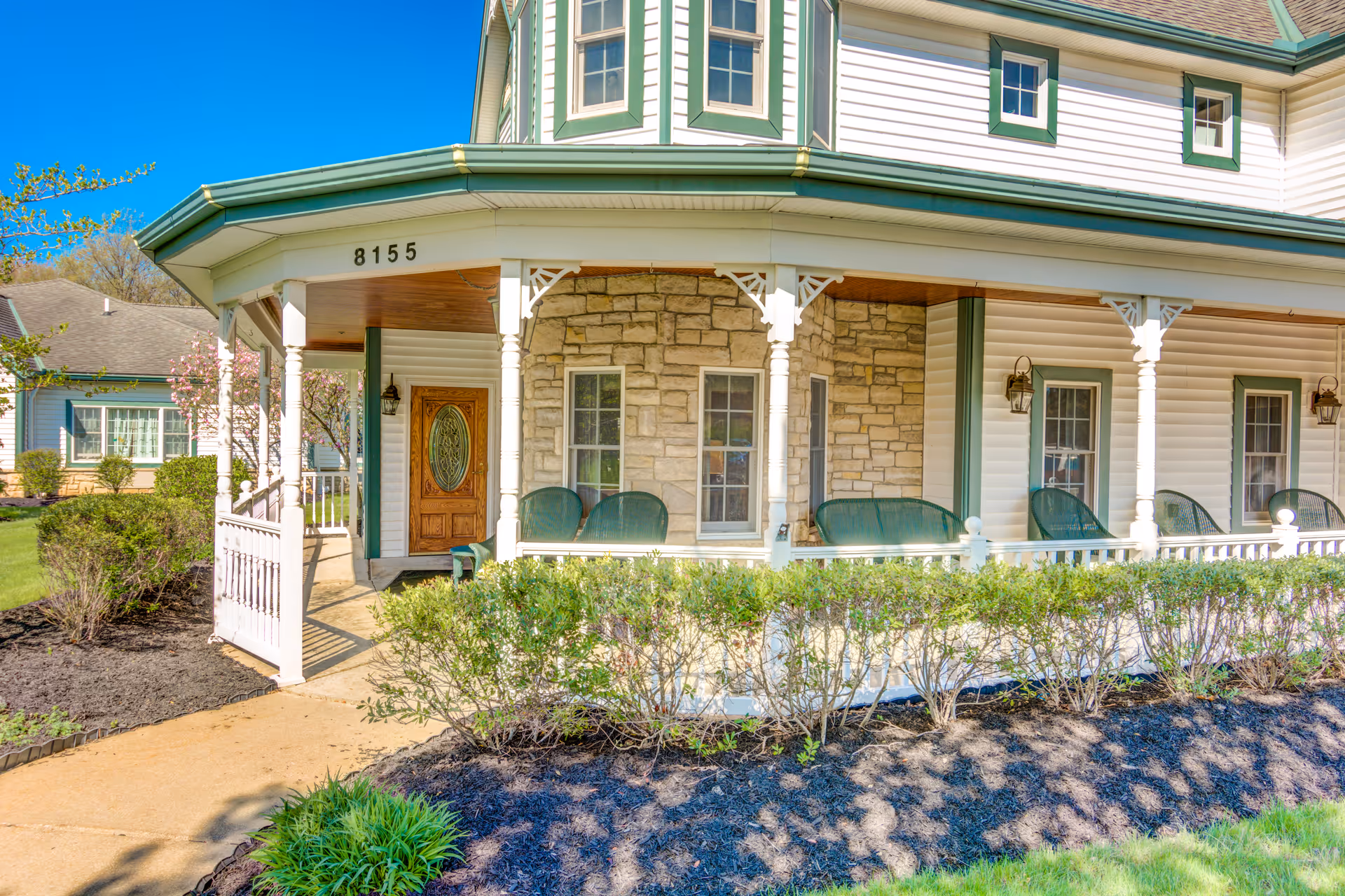 Exterior view of a senior living facility named Symphony at Mentor showing a wrap-around porch with green chairs, white railings, and a wooden front door with decorative glass. The building has white siding with green trim and stone accents, surrounded by landscaped bushes and a walkway.