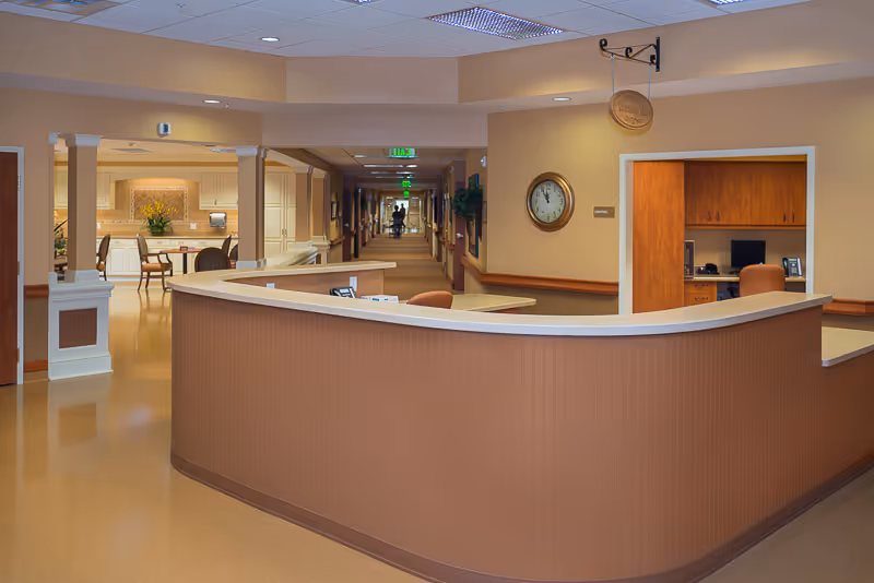 Reception desk area in a senior living facility with a curved counter, office space behind it, a clock on the wall, and a long hallway extending into the distance with exit signs and some furniture visible.