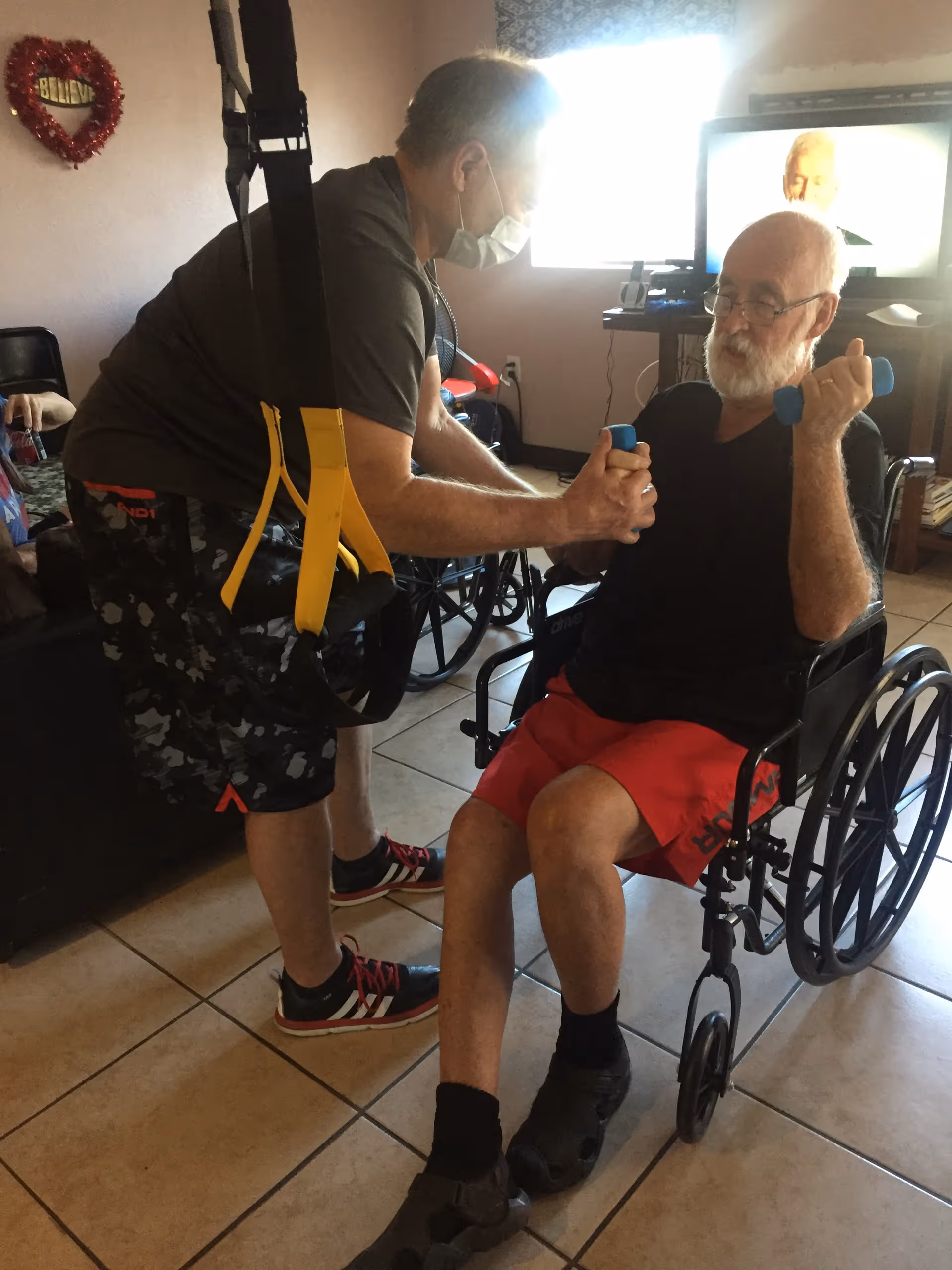 An elderly man in a wheelchair lifting small blue dumbbells while a caregiver wearing a face mask assists him with the exercise in a room with tiled flooring and a TV in the background.