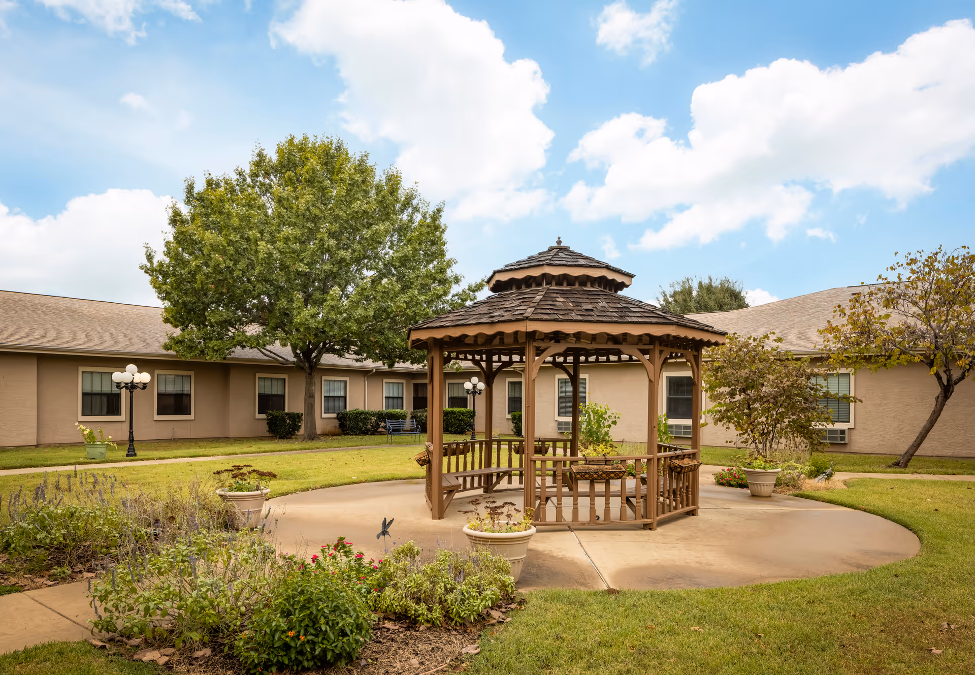 Outdoor courtyard area at a senior living facility featuring a wooden gazebo surrounded by a circular concrete path, green grass, potted plants, trees, and a beige building with multiple windows in the background under a partly cloudy sky.