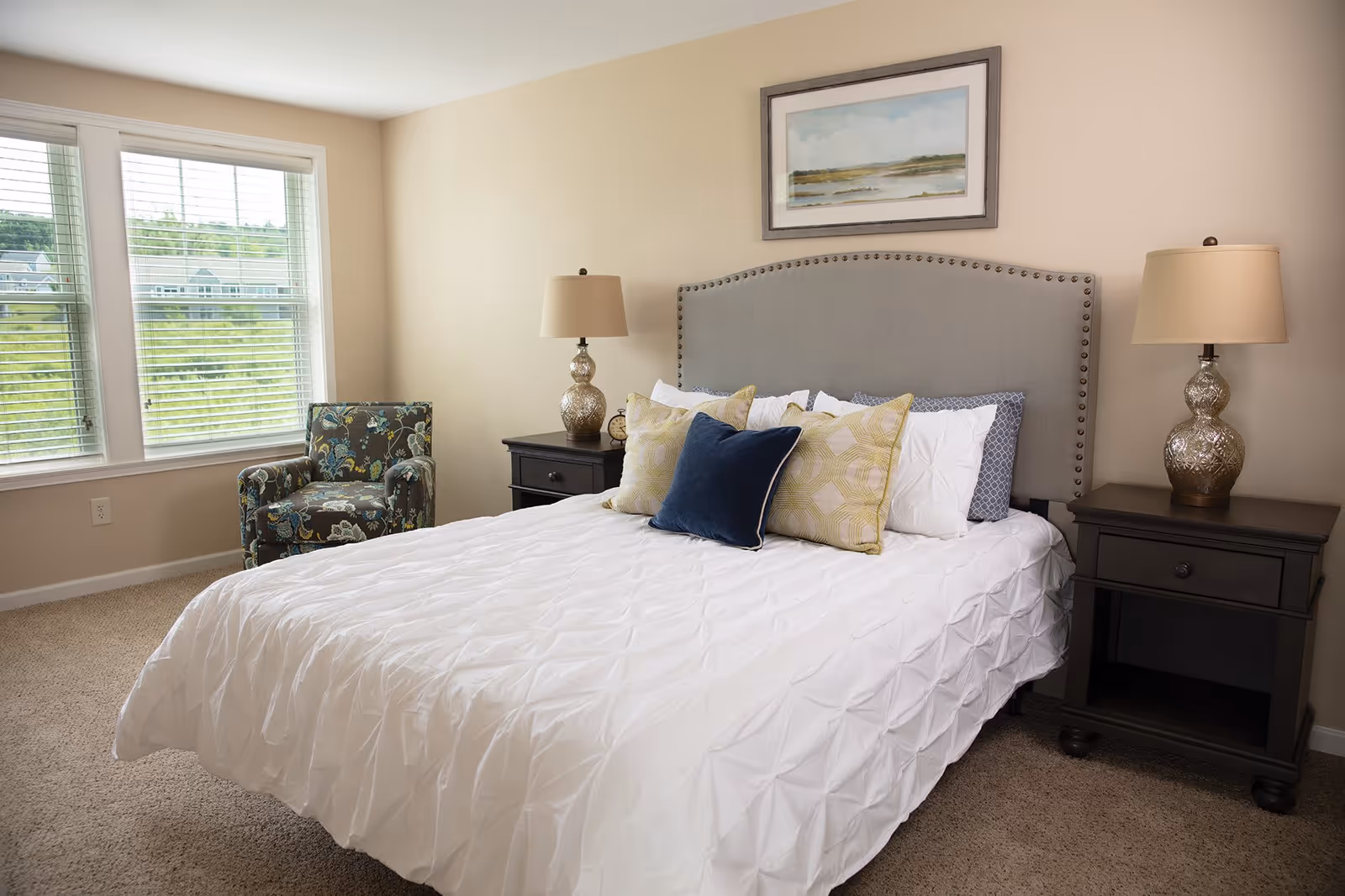 Sunlit bedroom with a bed dressed in white linens, an upholstered headboard, two bedside tables with lamps, a patterned armchair, and large windows.