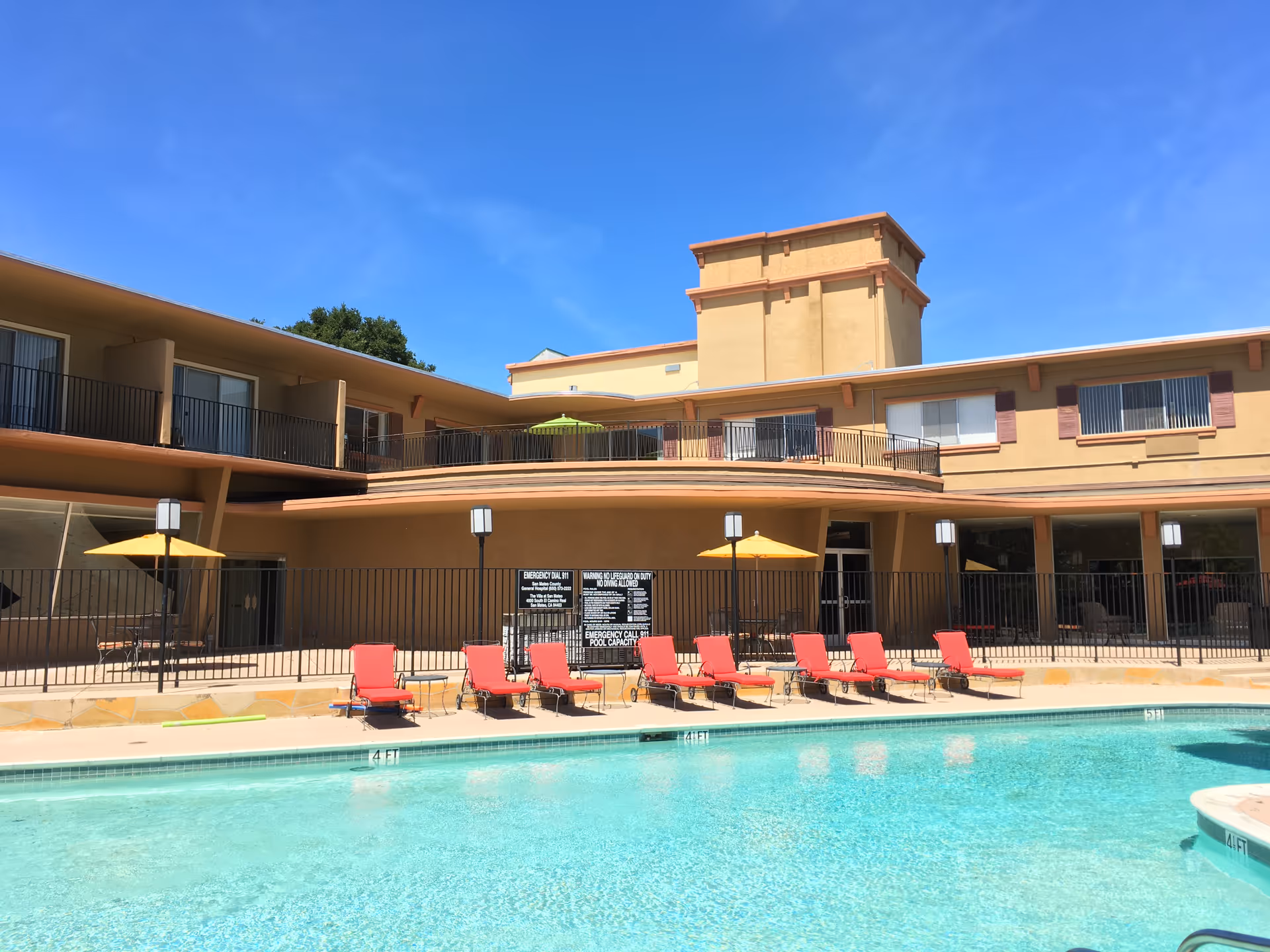 Outdoor swimming pool area at Villa at San Mateo with clear blue water, red lounge chairs lined up along the poolside, yellow umbrellas, and a two-story building in the background under a bright blue sky.