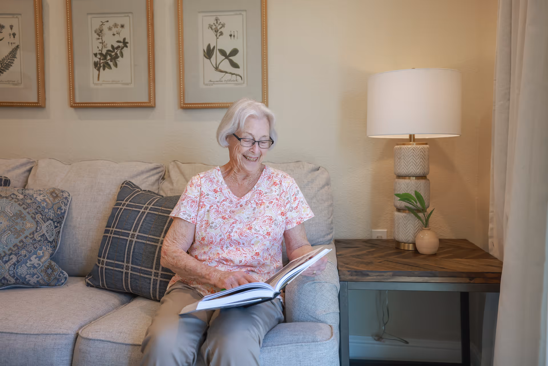 An elderly woman sits on a sofa smiling while reading a book in a cozy living room with framed botanical prints and a side table lamp.