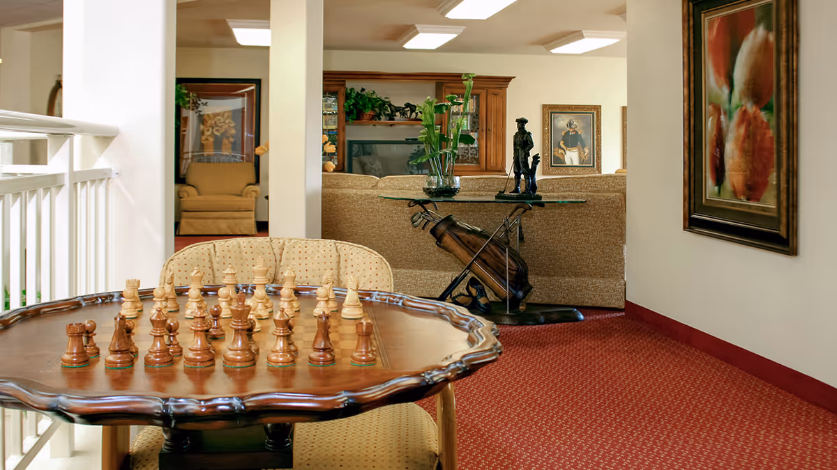 Interior view of a senior living facility lounge area with a wooden chess table and chairs in the foreground. In the background, there is a beige sofa, a glass table with decorative items, framed artwork on the walls, and a cozy armchair. The room has red carpet flooring and soft lighting.