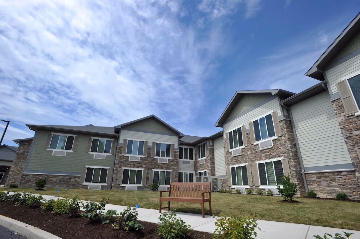 Exterior view of a two-story assisted living facility building with stone and siding facade under a partly cloudy blue sky. There is a wooden bench on a sidewalk in front of the building, surrounded by grass and small bushes.