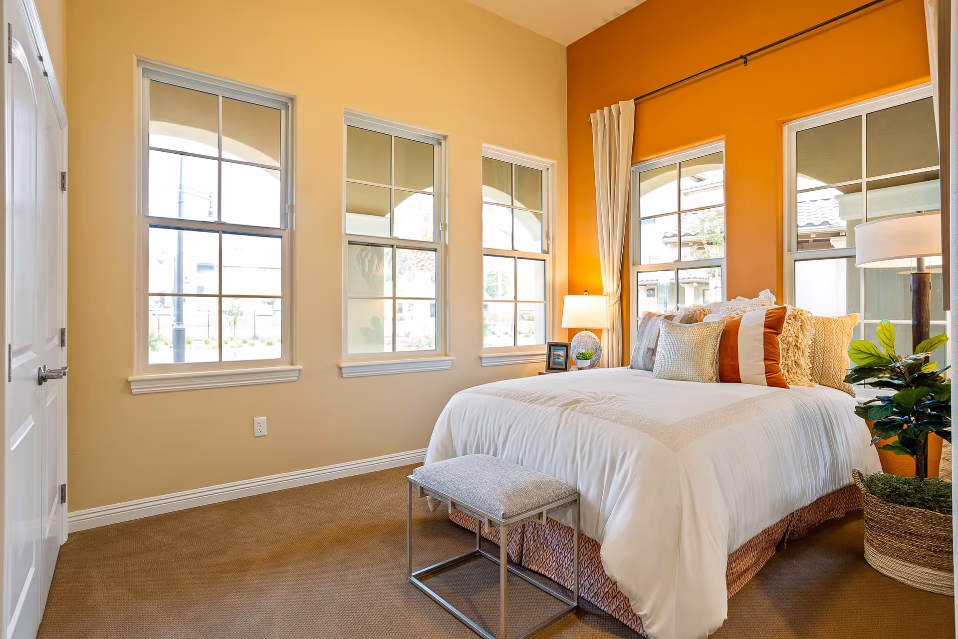A bright bedroom with a large bed covered in white bedding and multiple decorative pillows. The room features three large windows with white frames, beige walls with one accent wall painted orange, a small bench at the foot of the bed, a bedside table with a lamp and picture frame, and a potted plant in a woven basket on the floor.