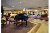 Spacious interior common area with a black grand piano and bench in the center, surrounded by comfortable seating including sofas and armchairs. The room features carpeted and tiled flooring, warm lighting, and decorative elements such as plants and framed pictures on the walls.